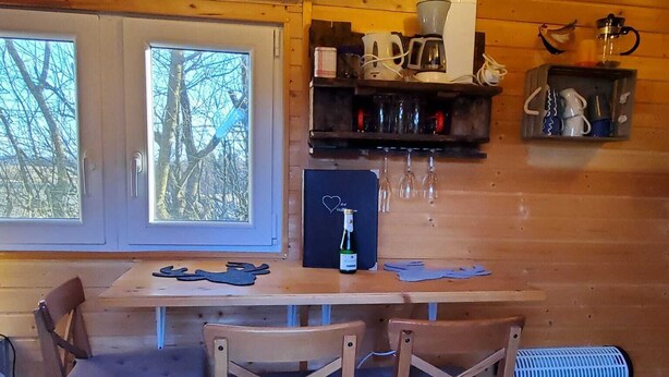 A wooden cabin interior with a dining table, chairs, a refrigerator, and a window with a view of trees. - Gemütlicher Schäferwagen in der Eifel (Nettersheim)