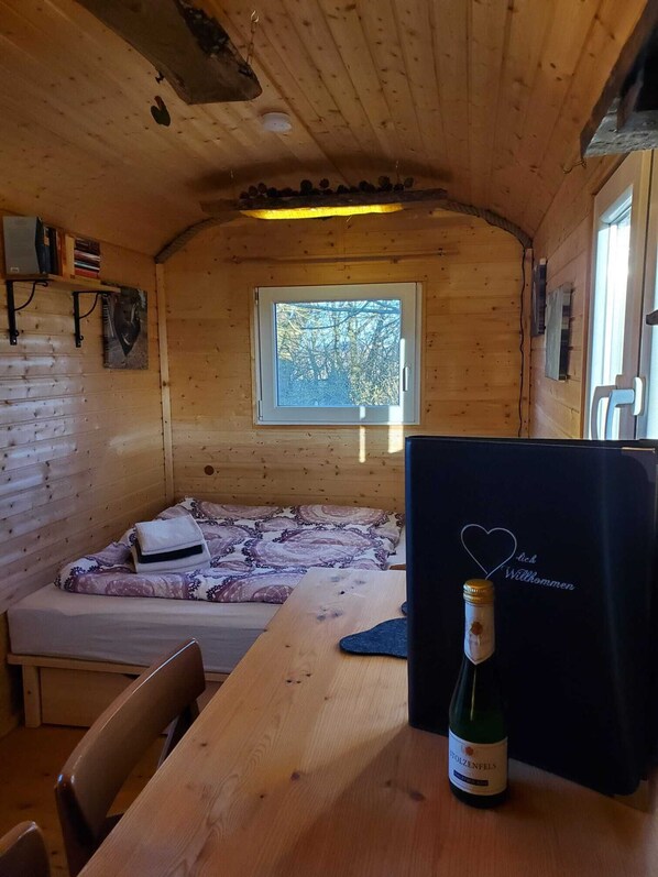 Acogedora habitación tipo cabaña con interior de madera, una cama con ropa de cama estampada, una pequeña mesa con una botella de vino y una ventana con vistas a árboles.