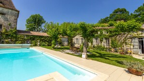 Une piscine dotée d’une terrasse carrelée, entourée d’une végétation luxuriante et de bâtiments historiques en pierre.