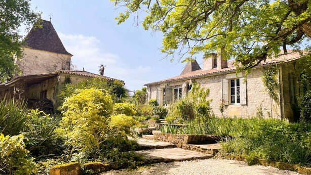 Un bâtiment rustique en pierre, doté d’un toit en tuiles, entouré d’une végétation luxuriante et bordé d’un chemin de gravier.