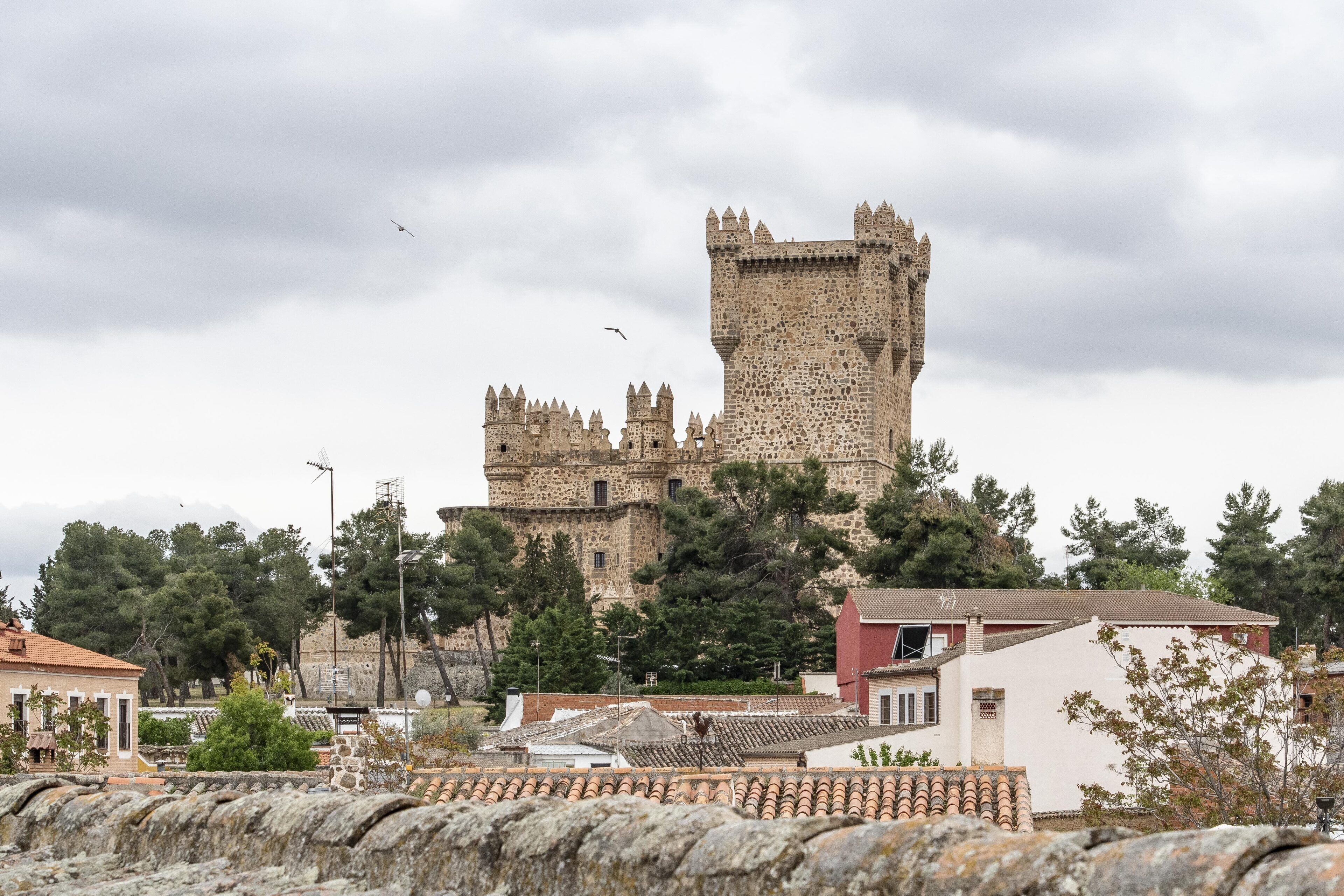A historic stone castle with a prominent tower, surrounded by modern buildings and trees.