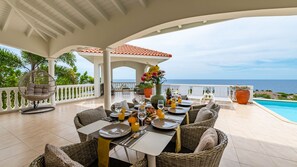 A dining area with a view of the sea, featuring wicker furniture, a hanging chair, and a table set for a meal. - Villa Summertime - Coral Estate Resort (Coral Estate)