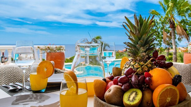 Een ontbijtsetting met een fruitschaal, glazen jus d'orange en een waterkan, met uitzicht op een zwembad en palmbomen.