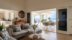 Een ruime woonkamer met een zithoek, een eettafel met stoelen en uitzicht op de zee door een open deur.