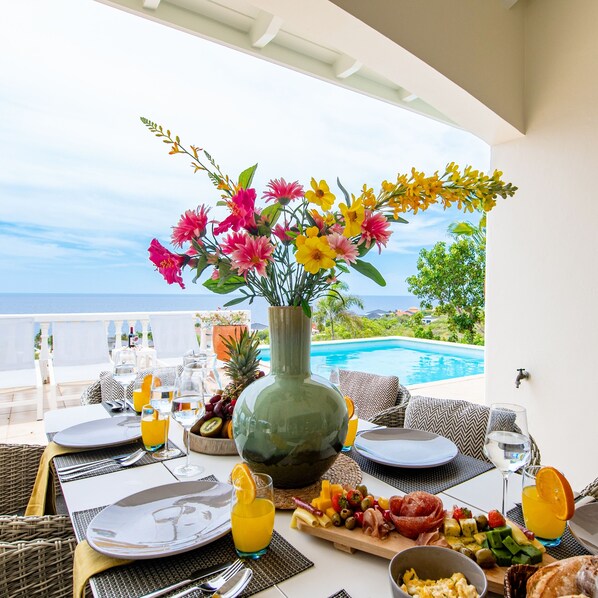 A dining table set with plates, cutlery, and a fruit platter, overlooking a pool and ocean view. - Villa Summertime - Coral Estate Resort (Coral Estate)