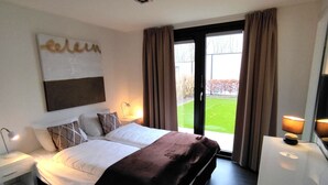 A modern hotel room with a large bed, a bedside table, a lamp, and a view of a green lawn through the window.
