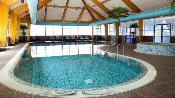 An indoor swimming pool with a tiled floor, a wooden ceiling, and a seating area with chairs.