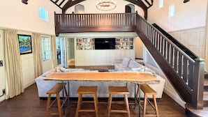 A spacious living area with a central table, multiple seating options, a staircase, and a bookshelf.