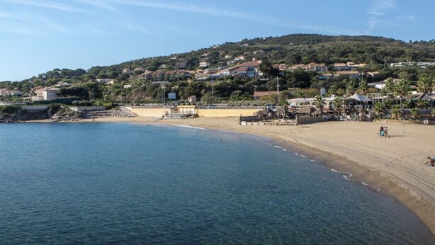 Eine Küstenlandschaft mit Sandstrand, einem kleinen Steg und einer Anhöhe mit Wohnhäusern.