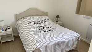 Ein ordentlich bezogenes Bett mit einem gestreiften Bettbezug und einem Kissen mit Schriftzug. Ein Nachttisch mit Lampe und ein kleiner Tisch ebenfalls mit Lampe. Der Raum hat Holzböden und weiße Wände.