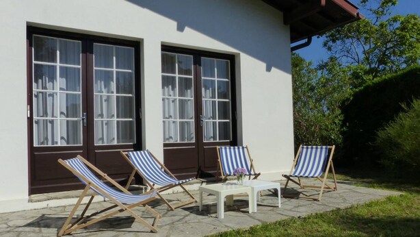 Una casa con dos ventanas grandes y un patio con dos sillones a rayas y una mesita.