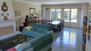 Una sala de estar con un sofá azul, una mesa de centro, una chimenea, una zona de comedor con mesa y sillas, y un ventanal con cortinas.