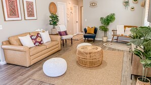 Een woonkamer met een beige bank, twee fauteuils, een salontafel en een groot, rond rieten pouf. Er liggen twee tapijten met een lijst, een potplant en een wandplank met decoratieve objecten.