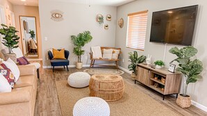 Een moderne woonkamer met een bank, poefjes, een televisie en decoratieve planten.