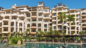 A hotel with multiple balconies, a swimming pool with lounge chairs, and palm trees. - Located on the pristine Medano beach (Cabo San Lucas)