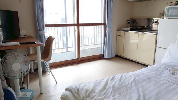 A hotel room with a bed, a desk with a chair, a fan, a kitchenette, and a large window with curtains. - Hokusei Building 41 (Sapporo)