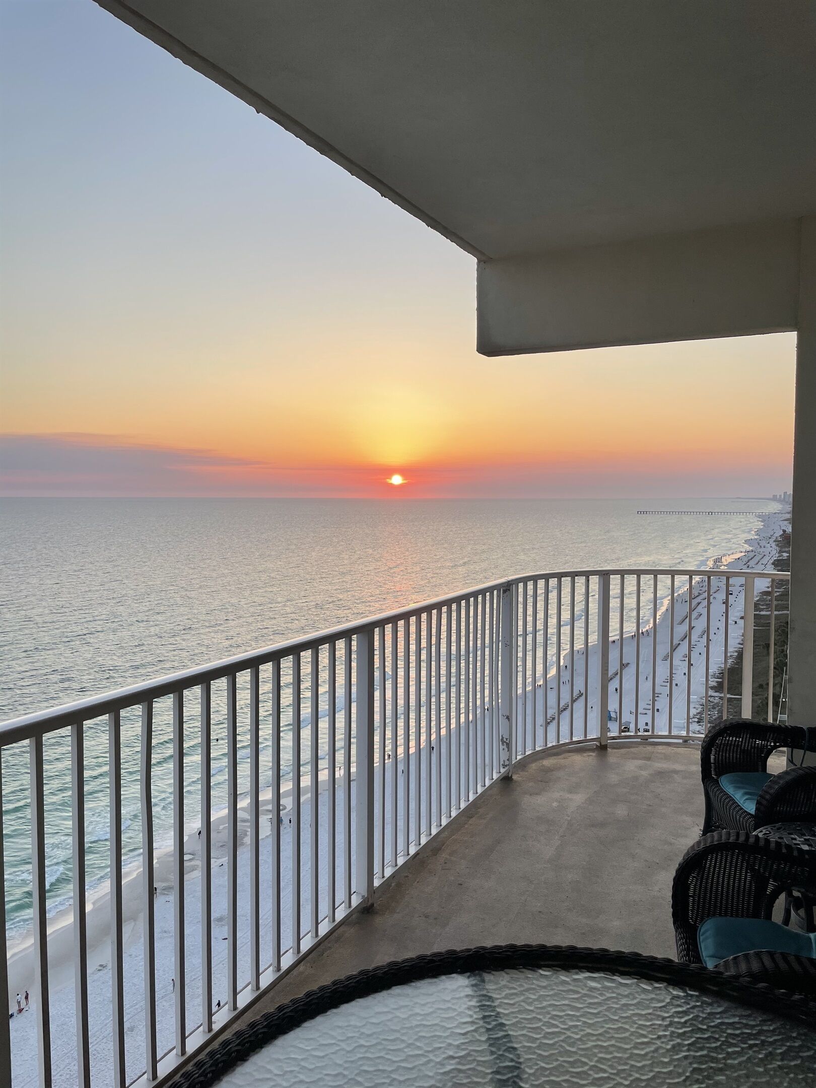 Een balkon met uitzicht op de oceaan tijdens de zonsondergang, met een witte balustrade en een rieten stoel.