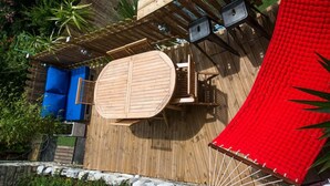 Una terraza de madera con una mesa redonda y sillas, un dosel rojo y una piscina azul.