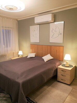 Un dormitorio con una cama, dos mesitas de noche con lámparas, un aire acondicionado y dos obras de arte colgadas en la pared.