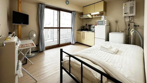 A hotel room with a bed, a desk with a chair, a kitchenette, and a large window. - Hokusei Building 42 (Sapporo)