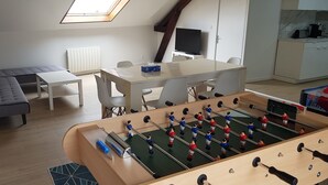 Une pièce comprenant une table de baby-foot, un canapé, une table à manger avec des chaises, un téléviseur et un coin cuisine.