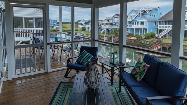 Ein überdachter Außenbereich mit Holzboden, einem Deckenventilator, einem blauen Sofa, einem Tisch, Stühlen und Blick auf Häuser über dem Wasser.
