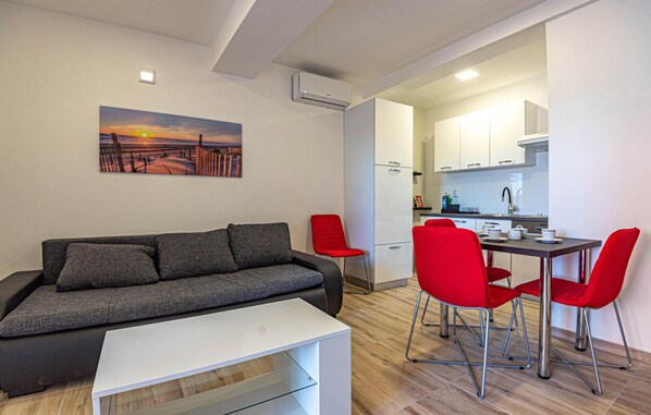 Ein modernes Wohnzimmer mit einem grauen Sofa, einem kleinen weißen Tisch und roten Stühlen. Im Hintergrund ist eine Küche mit weißen Schränken und einem Esstisch erkennbar.