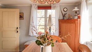 Et spiseområde med et træbord, en vase med blomster, et vindue med gardiner, et træskab og et ur på væggen.