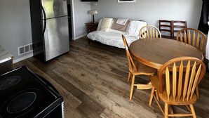Ett kompakt boendeutrymme med ett litet kök, en matplats med trästolar, en säng med kuddar och en platt-TV som är monterad på väggen.