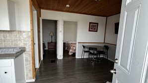 A spacious room with a wooden ceiling, a kitchen area with a tiled backsplash, and a dining table with chairs. - Dock Life Beach 1 - Cottage 3, Riverside 2 bedroom, 3 min walk to Beach 1 (Wasaga Beach)