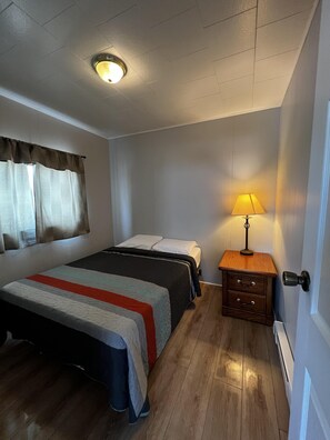A bedroom with a bed, a nightstand with a lamp, and a window with curtains. - Dock Life Beach 1 - Cottage 4, Riverside 2 bedroom, 3 min walk to Beach 1 (Wasaga Beach)