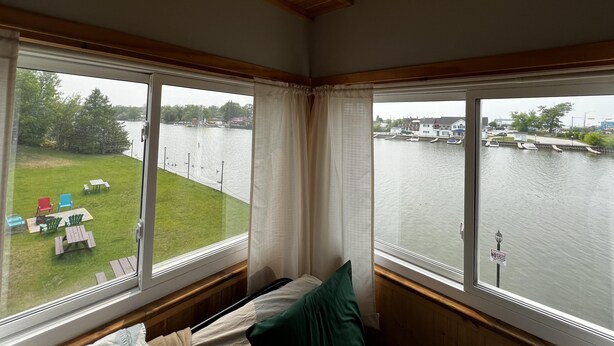 Ein Zimmer mit Seeblick, eine Wiese mit Stühlen und Tischen davor sowie ein Bett mit einem Kopfkissen.