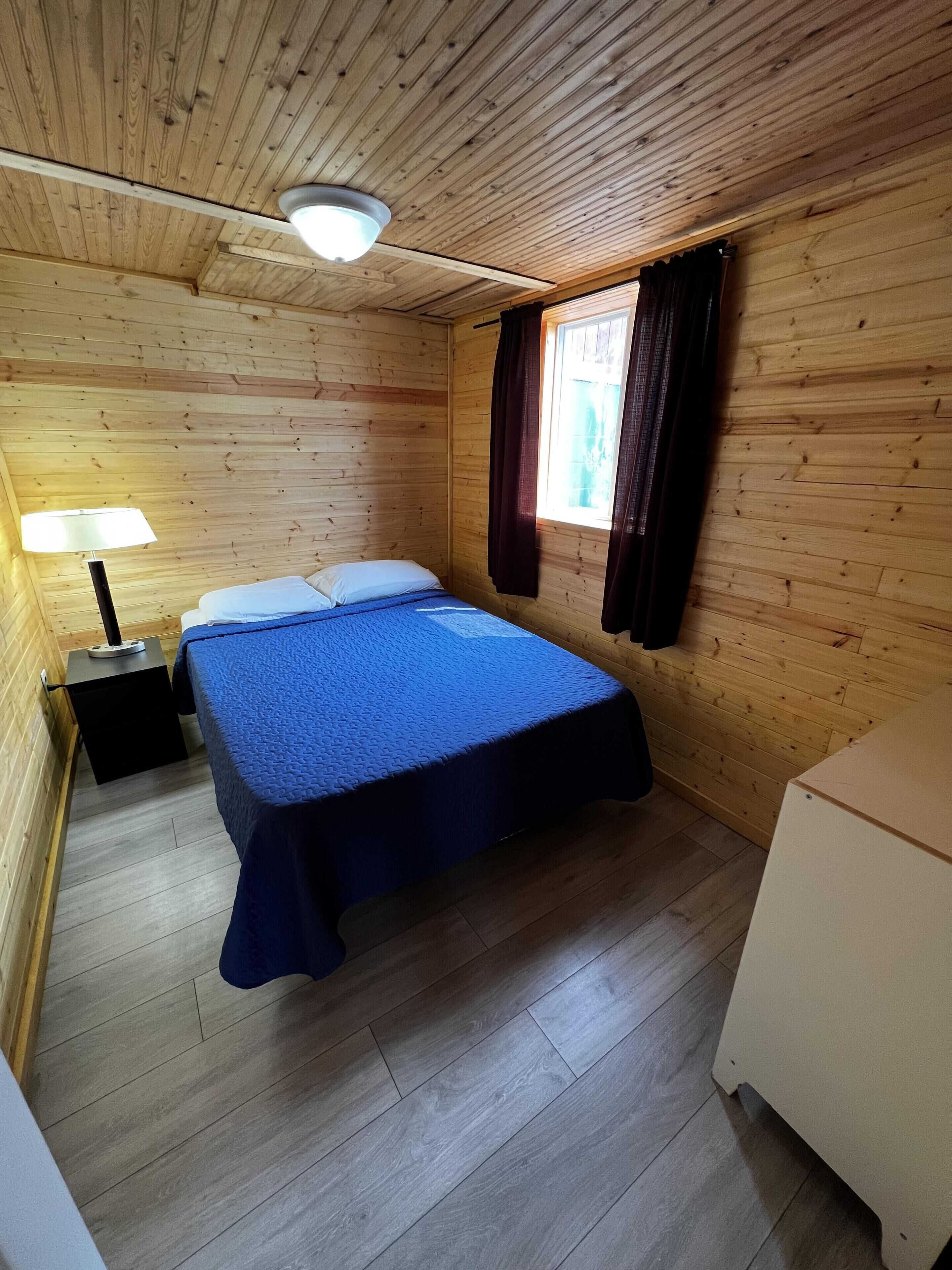 Een kamer met houten lambrisering, een bed, nachtkastje, lamp en een raam met gordijnen.