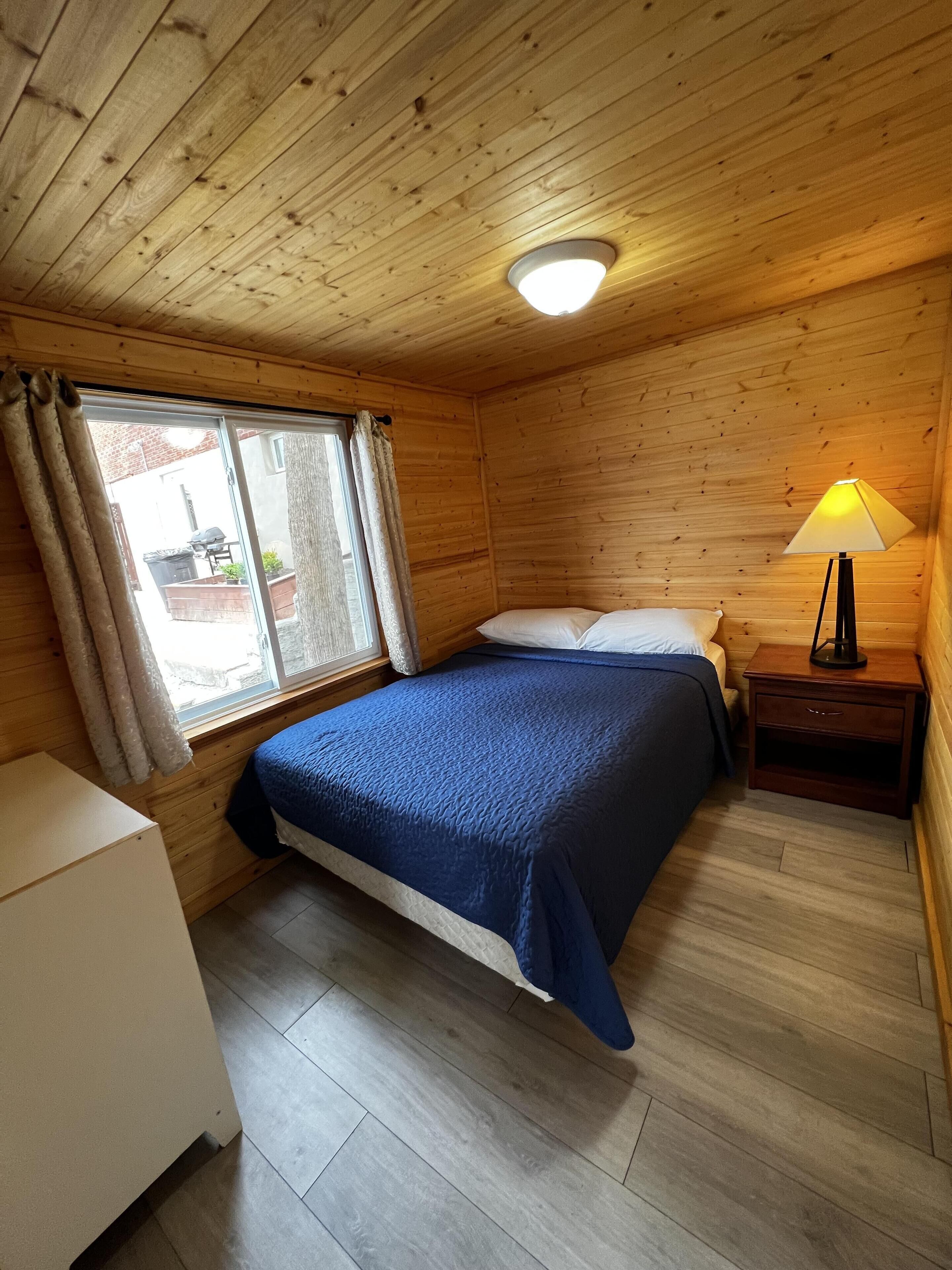 Une pièce de style cabane en bois, avec un lit, une table de chevet dotée d’une lampe, une fenêtre avec des rideaux et une petite armoire blanche.