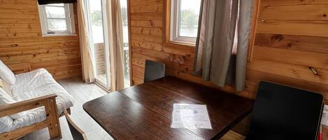 A wooden cabin with a dining table, two chairs, a sofa, and a window overlooking a natural landscape.