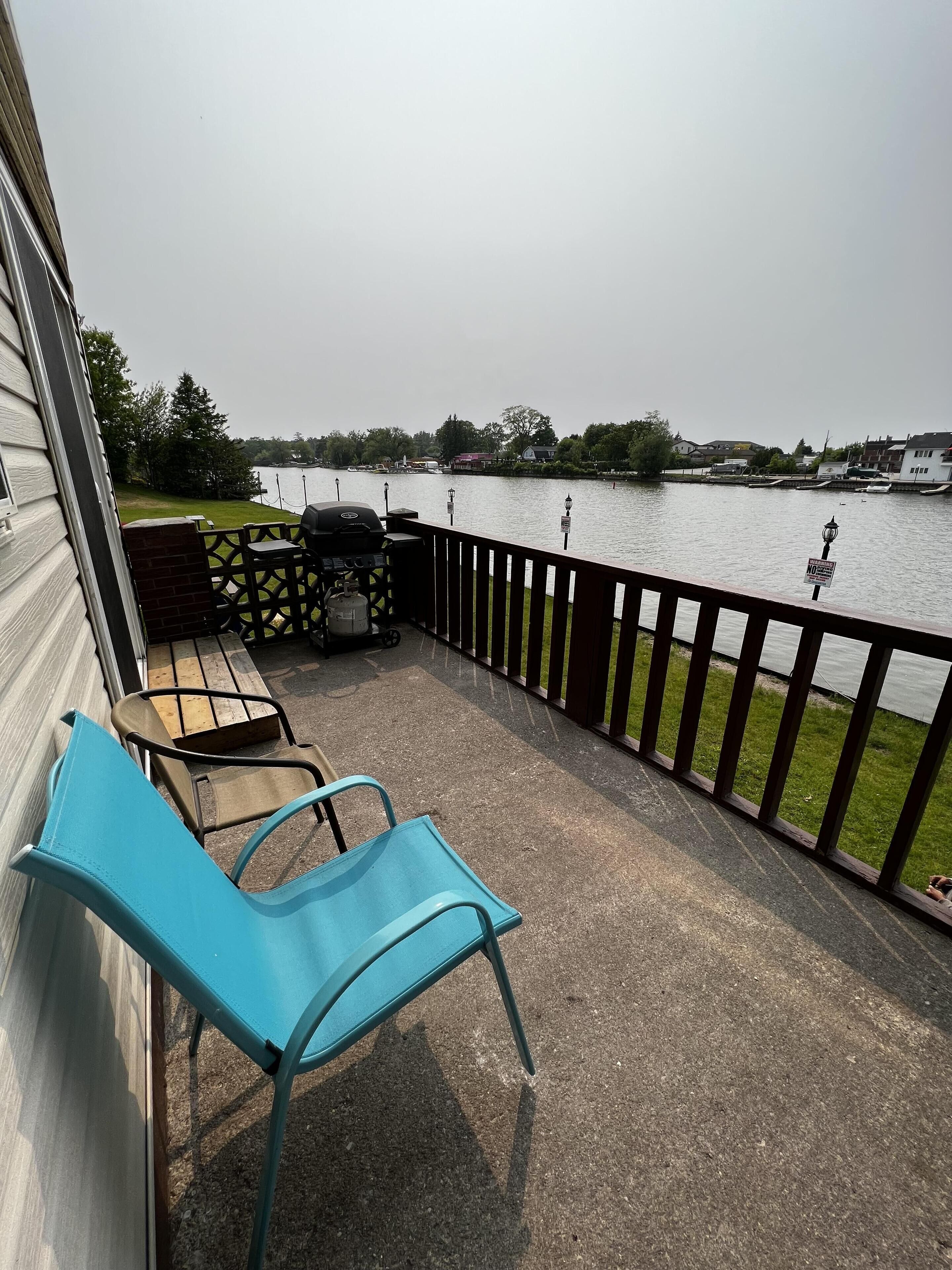 A balcony with a view of a lake, a grill, and a few chairs.