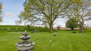 A well-maintained lawn with a stone fountain, large trees, and a distant view of a field.