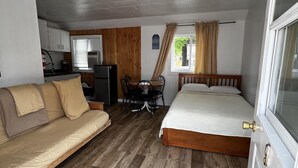 A compact room with a wooden floor, a sofa, a bed, a small table with chairs, and a kitchenette area. - Dock Life Beach 1 - Cottage 9, Riverside Property, Studio, 3 min walk to Beach 1 (Wasaga Beach)