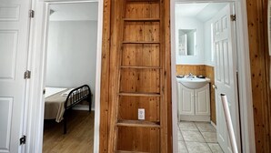 Ett sovrum på entresolplan med en trästege som leder upp till en högre nivå, en säng och ett badrum med handfat och spegel.