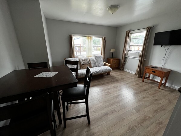 A room with a wooden dining table, two chairs, a sofa, a small bedside table with a lamp, and a TV mounted on the wall. - Dock Life Beach 1 - Unit 16, Riverside Property, 2 Bdrm, 3 min walk to Beach 1 (Wasaga Beach)