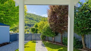 A covered patio with a gravel path leading to a grassy area, surrounded by trees and a fence. - Duff's Cottage-fabulous location-comfort +style (Arrowtown)