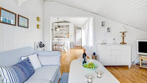 Una habitación luminosa, con paredes blancas y techo de madera, un sofá con cojines estampados, una mesa de comedor con una botella de vino y un bol de uvas, y un televisor sobre un mueble.
