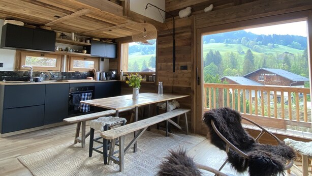 A rustic cabin interior with a wooden ceiling, a kitchen area, a dining table with benches, and a view of a mountainous landscape through large windows.