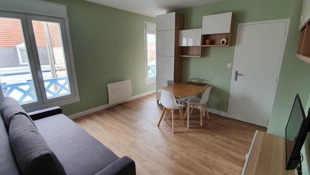 Une pièce de taille modeste avec un plancher en bois, un canapé gris, une table à manger avec des chaises et un téléviseur fixé au mur.