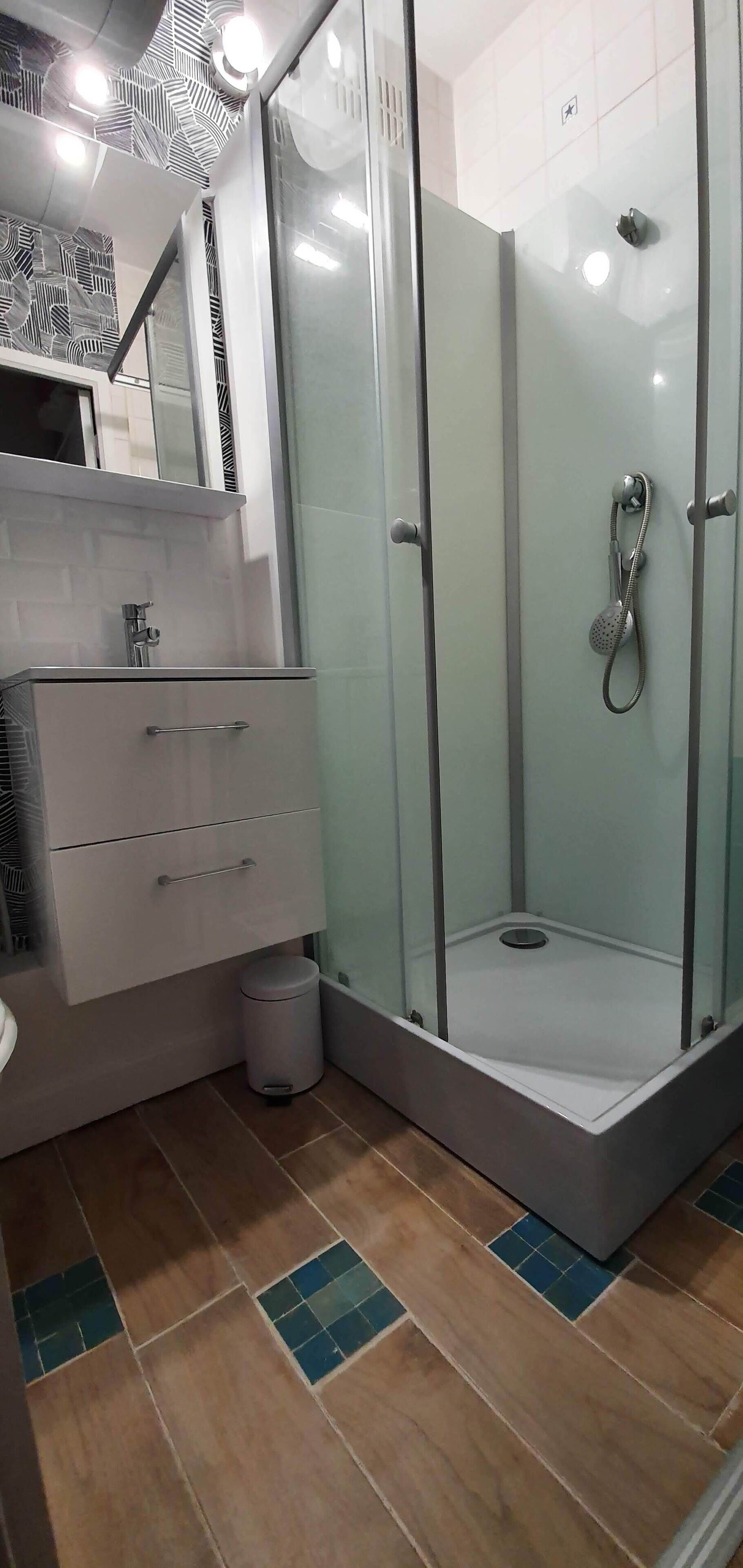 Uma casa de banho moderna com chuveiro em cabine de vidro, um lavatório branco com gavetas e um chão em mosaico com padrão geométrico.