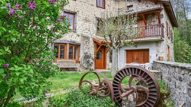 Une maison en pierre avec un balcon en bois, une porte en bois et une grande roue à eau dans le jardin.
