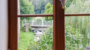 Une vue à travers une fenêtre sur un pont de pierre enjambant un ruisseau, le tout entouré d’une végétation luxuriante.