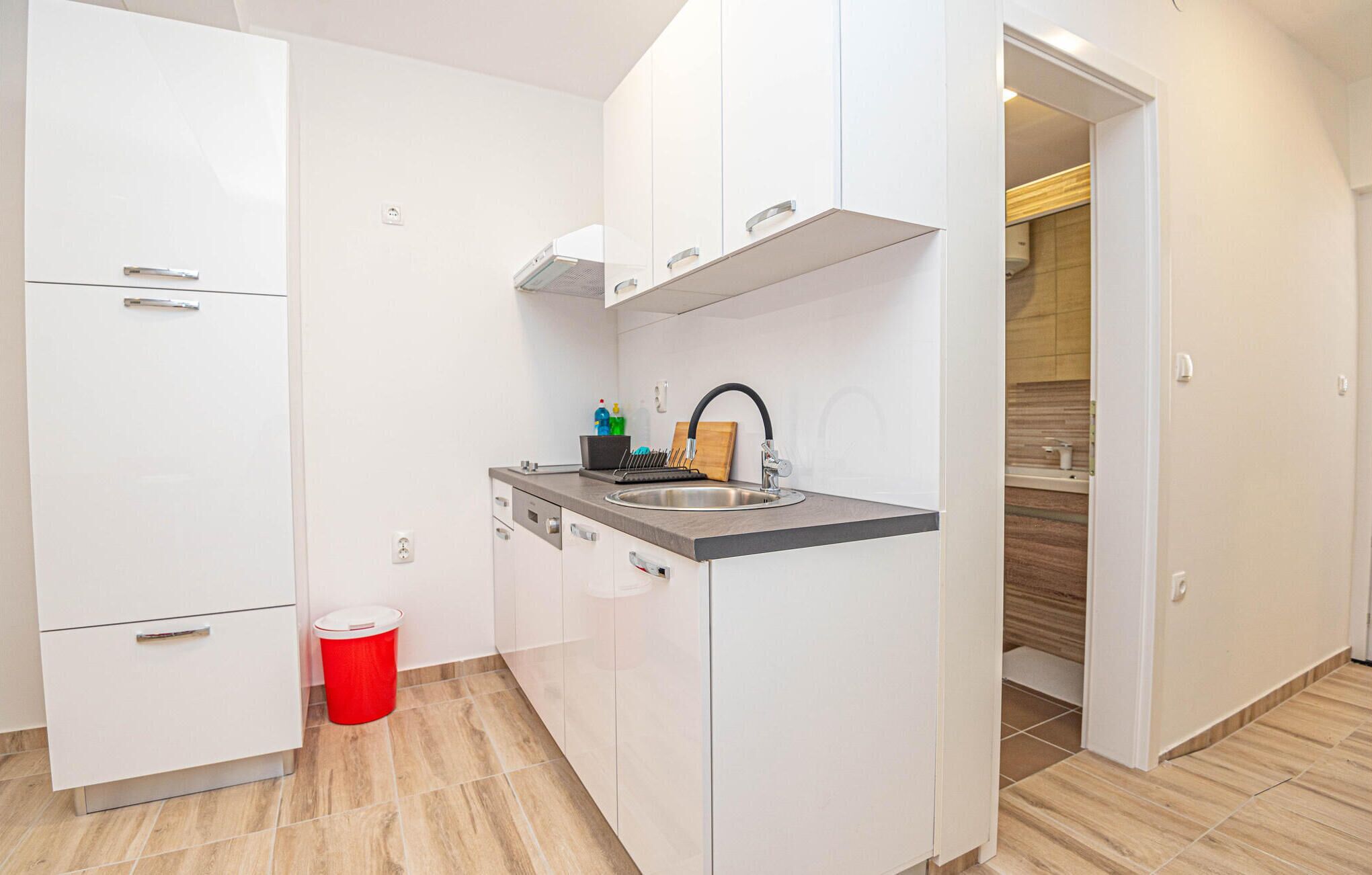 A compact kitchen with white cabinets, a stainless steel sink, and a red trash bin.