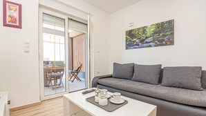 Ein modernes Wohnzimmer mit einem grauen Sofa, einem Couchtisch mit Tassen und einer Schale, einem Bild von einem Waldbach und einer Glastür, die auf einen Balkon mit Esstisch und Stühlen führt.