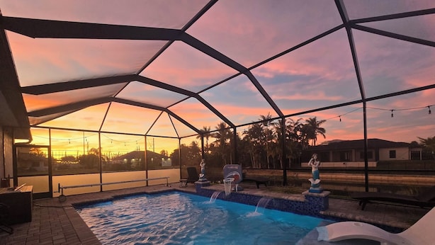 A swimming pool with a waterfall feature, surrounded by a covered area with a view of palm trees and a sunset. - Gorgeous build on fresh water canal - very private-heated pool (Cape Coral)